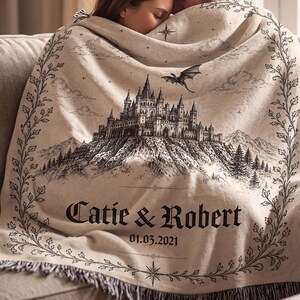 Op de afbeelding: Beige deken met een zwart-wit illustratie van een kasteel, een draak, bergen en bomen. De deken heeft de namen "Catie & Robert" en de datum "01.03.2021" erop gedrukt, met een decoratieve rand.