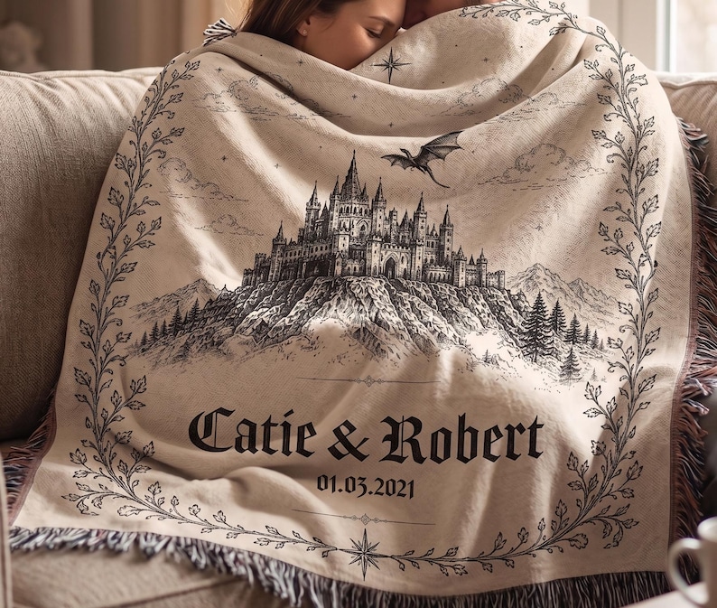 Op de afbeelding: Beige deken met een zwart-wit illustratie van een kasteel, een draak, bergen en bomen. De deken heeft de namen "Catie & Robert" en de datum "01.03.2021" erop gedrukt, met een decoratieve rand.