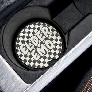 Puede incluir: Posavasos de coche a cuadros blanco y negro con el texto "ELDER EMO" en blanco. El posavasos está en un portavasos de coche.