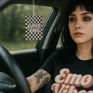 Pode incluir: Uma camiseta preta com as palavras "Emo Vibes" em uma fonte retrô. Um purificador de ar xadrez com as palavras "Elder Emo" pendurado no espelho retrovisor. A pessoa no carro tem tatuagens nos braços.