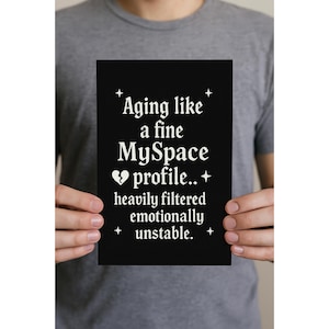 May include: A black rectangular print with white text that reads "Aging like a fine MySpace profile.. heavily filtered emotionally unstable." The print is held by a person wearing a gray t-shirt.