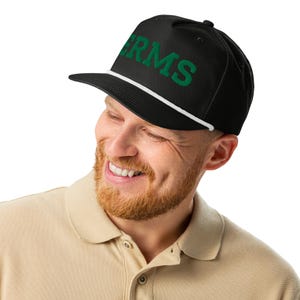 Puede incluir: Gorra negra de ala plana con la palabra "RMS" en letras verdes. La gorra tiene un detalle de cuerda blanca a lo largo del borde. La persona de la imagen lleva un polo beige.