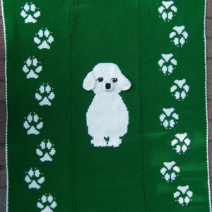 Può includere: Una coperta di maglia verde con un barboncino bianco al centro e impronte di zampe bianche sui lati.