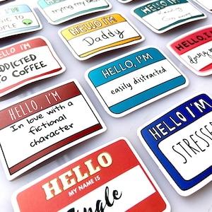 Può includere: Una collezione di adesivi con etichette con varie frasi. Gli adesivi hanno uno sfondo bianco con bordi colorati e testo come "Hello, I'm Daddy", "Hello, I'm easily distracted" e "Hello, my name is Single".