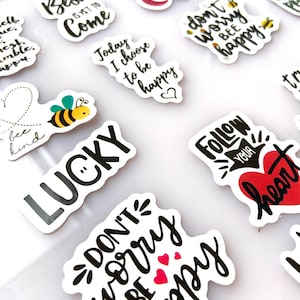 Puede incluir: Una colección de pegatinas blancas con letras negras y varios diseños. Las pegatinas presentan frases motivacionales como "The Best is Yet to Come", "Don't Worry, Be Happy" y "Follow Your Heart". Algunas pegatinas incluyen pequeñas ilustraciones.
