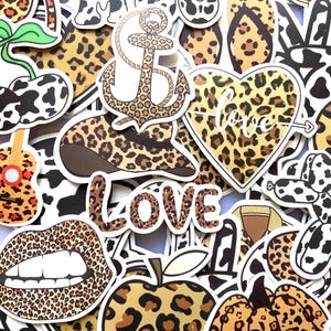 Op de afbeelding: Een verzameling diverse stickers met luipaard- en koeienprintpatronen. De ontwerpen omvatten een anker, een hart met het woord love, een gitaar, lippen, een appel, een pompoen en diverse vormen, allemaal voorzien van zwarte omlijningen en witte randen.