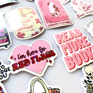 Könnte beinhalten: Eine Sammlung farbenfroher Aufkleber mit verschiedenen Designs und Texten. Aufkleber enthalten Sätze wie „Read More Books“, „I'm Here for Red Flags“ und „Friends to Lovers“. Weitere Aufkleber zeigen Buchgrafiken und Zitate.