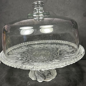 May include: A clear glass cake stand with a domed lid. The stand features a decorative, raised floral pattern on the base and a beaded edge. The lid has a knob handle. The stand is designed to display and protect baked goods.