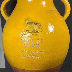 May include: Yellow ceramic olive oil jar with two handles. The jar has a brown base and features the words "Olio Di Oliva Extra Vergine" in white lettering. A gold emblem is also visible.