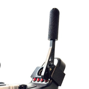 Puede incluir: Palanca de cambios de carreras negra con una empuñadura negra texturizada y cinco botones rojos. La palanca de cambios está montada en una base negra.