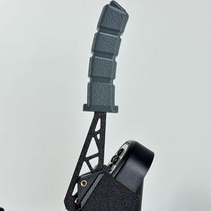 May include: A black and gray gaming controller with a textured grip. The controller features a lever with a textured handle and a black base with buttons and a velcro strap. The design is modern and sleek.