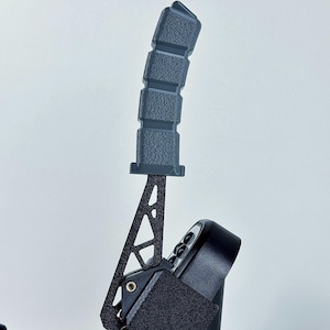 Puede incluir: Un freno de mano negro para simulador de carreras con un agarre texturizado. El freno de mano tiene un marco de metal negro y una correa de velcro. El agarre está hecho de un material gris oscuro.