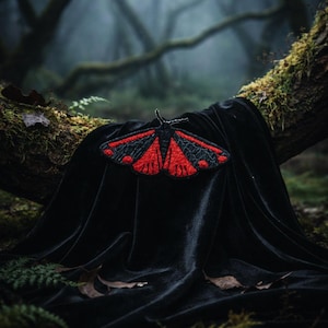 May include: A black velvet cloth draped over a moss-covered tree branch, with a red and black moth-shaped patch. The patch has detailed embroidery, set against a blurred forest backdrop.