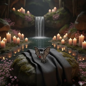 Peut inclure: Un écusson en forme de chauve-souris marron avec un cœur rouge est présenté sur un tissu gris foncé drapé sur une pierre. L'arrière-plan présente une cascade, des bougies et des fleurs roses, créant une scène mystique.