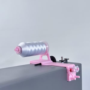 Könnte beinhalten: Ein rosa Garnrollenhalter mit einer silbernen Garnrolle. Der Halter ist an einer grauen Oberfläche befestigt. Der Faden kommt aus der Spule.