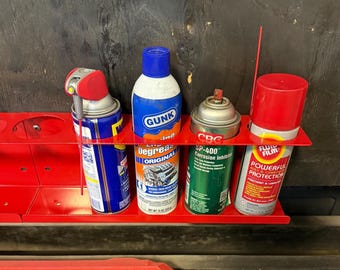 Puede incluir: Un estante metálico rojo sostiene cuatro botes de productos automotrices. Los botes incluyen Gunk Degreaser, CRC SP-400 Corrosion Inhibitor y Fluid Film Powerful Protection. El estante está montado sobre una superficie oscura.