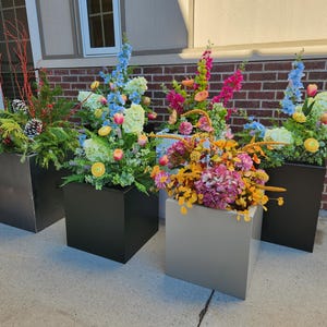 May include: Four square planters filled with colorful floral arrangements. The planters are in shades of gray and black. The flowers include blue, pink, yellow, and orange blooms, with greenery and red branches.