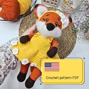 May include: A handmade crochet fox doll with orange, white, and black details, wearing a yellow dress adorned with white flower accents. The doll has a headband with white flowers. The image includes text that says "Crochet pattern PDF".