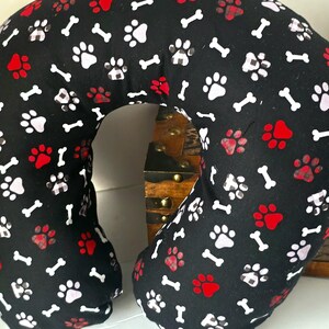 May include: A black travel pillow with a pattern of white bones, red paw prints, and white and red paw prints. The pillow is U-shaped, designed for neck support, and rests on a wooden surface.