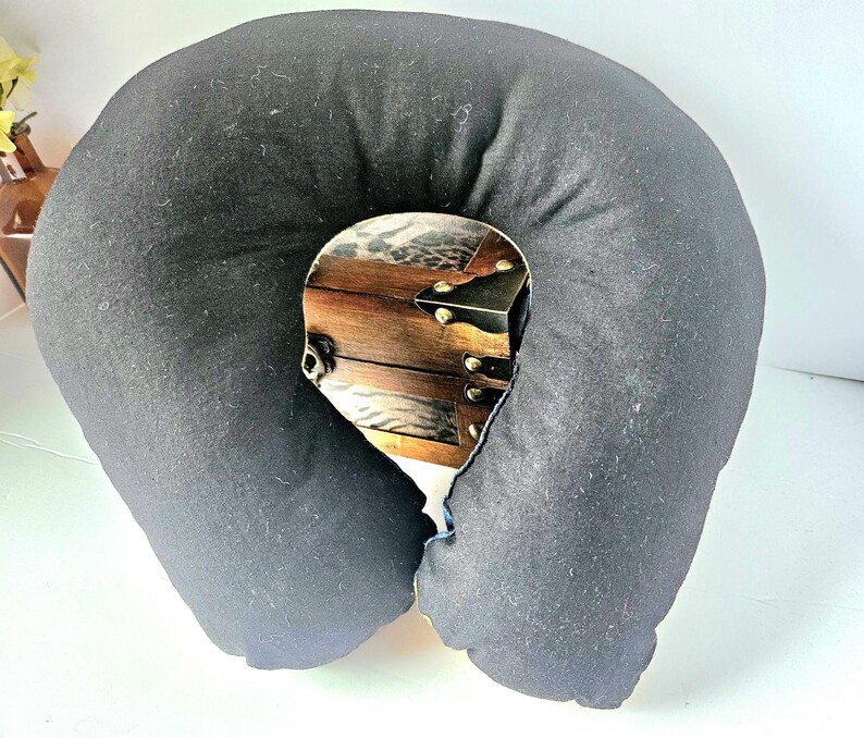 May include: A black travel pillow with a decorative wooden chest design in the center. The pillow is U-shaped and made of soft fabric. The chest design features brown wood, metal accents, and a leopard print.