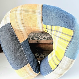 May include: A U-shaped travel pillow with a patchwork design. The pillow features denim, yellow plaid, and tan fabric sections. The pillow is designed for neck support and comfort.