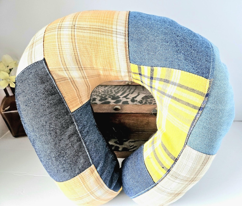 May include: A U-shaped travel pillow with a patchwork design. The pillow features denim, yellow plaid, and tan fabric sections. The pillow is designed for neck support and comfort.