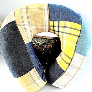 May include: A patchwork travel pillow with a U-shape design. The pillow features a variety of fabrics in shades of yellow, blue, and gray. The fabrics include plaid and solid textures. The pillow is designed for neck support and comfort.
