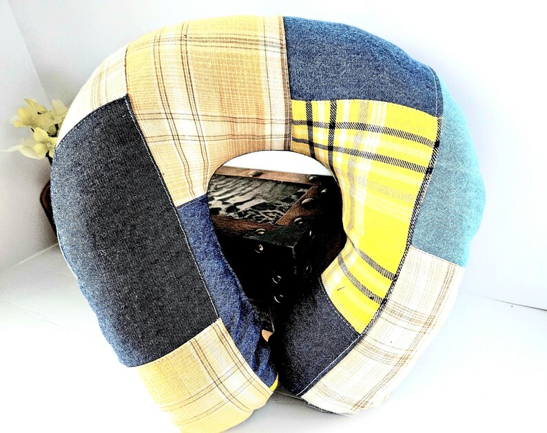 May include: A patchwork travel pillow with a U-shape design. The pillow features a variety of fabrics in shades of yellow, blue, and gray. The fabrics include plaid and solid textures. The pillow is designed for neck support and comfort.