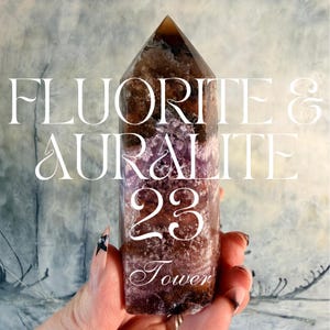 May include: A Fluorite & Auralite 23 tower crystal. The crystal is a mix of brown, purple, and white hues. The tower is held in a hand with a painted nail. The text "Fluorite & Auralite 23 Tower" is displayed on the image.