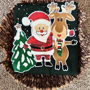 Puede incluir: Una camiseta verde oscuro con temática navideña que presenta a Papá Noel, un reno y un árbol de Navidad. Papá Noel lleva un traje y gorro rojos, y el reno tiene una bufanda verde y blanca. El árbol es verde con una estrella dorada.
