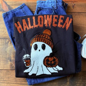 Puede incluir: Sudadera negra de Halloween con un simpático diseño de fantasma que lleva un gorro de punto y sostiene una calabaza y una bebida. La palabra "HALLOWEEN" está escrita en la parte superior con purpurina naranja. La sudadera se combina con vaqueros azules.