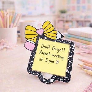 May include: A decorative memo holder shaped like a composition notebook page with a yellow sticky note. The note reads "Don't forget! Parent meeting at 3 pm ♡". The holder is adorned with a yellow pencil and bow design with a pink heart.