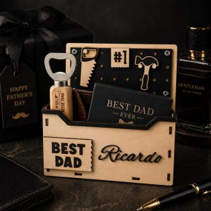 Puede incluir: Un organizador de escritorio de madera con las palabras "BEST DAD" y "Ricardo" grabadas en la parte delantera. Contiene un abrebotellas, una pequeña tarjeta que dice "BEST DAD - EVER" y recortes decorativos de madera. Una caja de regalo negra y un frasco de colonia están en el fondo.