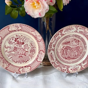 Peut inclure: Deux assiettes rondes en céramique ornées d'un motif de transfert rouge foncé sur fond crème. L'assiette de gauche présente un motif central de vase floral, tandis que celle de droite montre un paysage détaillé. Bordures à treillis complexes.