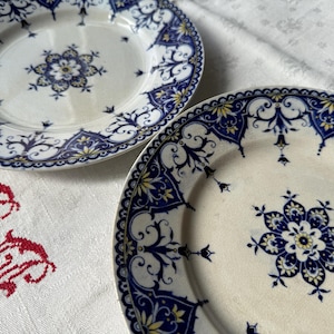 May include: Two white ceramic plates with intricate blue and yellow floral designs. The plates feature a central floral motif and a detailed border. The plates are placed on a white tablecloth with red embroidered details.