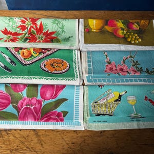 Op de afbeelding: Een verzameling vintage linnen theedoeken met diverse ontwerpen. De theedoeken tonen bloempatronen, stillevens met fruit en illustraties met keukenthema's. Kleuren zijn onder meer groen, blauw, roze en rood. De theedoeken zijn opgevouwen en op een houten oppervlak geplaatst.