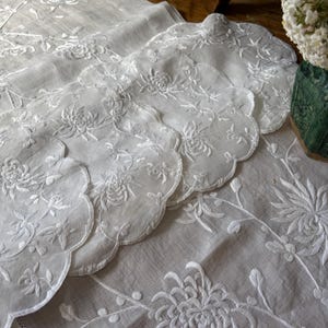 May include: A set of white fabric doilies and a matching runner featuring intricate white floral embroidery. The pieces have scalloped edges and are decorated with detailed chrysanthemum-like flower patterns stitched in white thread.