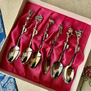 May include: A set of six silver-toned metal spoons arranged in a light-colored box with a bright pink satin lining. Each spoon features a handle shaped like a textured tree branch with a unique animal or bird figure perched at the very top end.