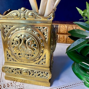 May include: A rectangular brass container featuring intricate openwork filigree patterns and a central circular medallion with symmetrical scrollwork. The metallic gold-toned vessel has a decorative top border and a solid base, designed to hold items.