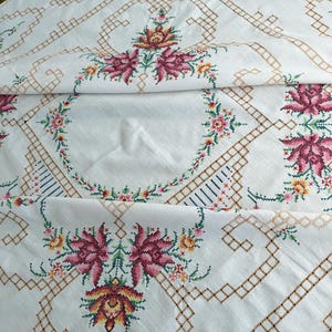 May include: A white tablecloth featuring intricate cross-stitch embroidery in shades of deep pink, yellow, and green. The design includes floral motifs arranged in a repeating pattern, connected by geometric brown lattice lines across the fabric.