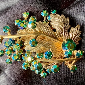 May include: A vintage gold-tone brooch with a leaf design and clusters of green and blue iridescent rhinestones. The brooch features a detailed leaf and branch design, with the rhinestones adding sparkle.