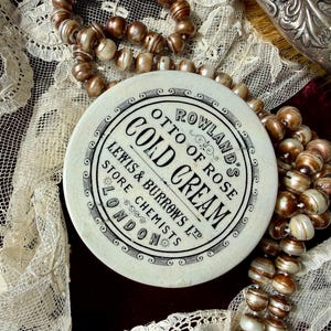 Op de afbeelding: Een rond, gebroken wit keramisch deksel met zwarte opdruk: ROWLAND'S OTTO OF ROSE COLD CREAM, LEWIS & BURROWS LTD, STORE CHEMISTS, LONDON. Het deksel is omringd door een streng iriserende, lichtbruine, bolvormige kralen.