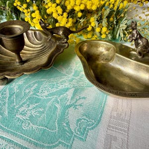 May include: Two brass decorative dishes with metallic animal figures. The left dish is a shell-shaped candle holder with a cat figure. The right dish is a divided tray with a kangaroo figure and the engraved text "Mann April 18th 1924".