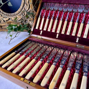 Könnte beinhalten: Ein hölzerner Besteckkasten mit einem Set aus versilberten Gabeln und Messern mit cremefarbenen Griffen. Das Innere ist mit tiefrotem Samt ausgekleidet. Die Messerklingen weisen detaillierte florale Gravuren auf und die Griffe tragen Monogramme.