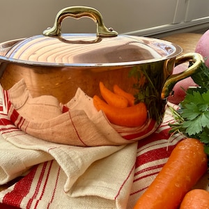 May include: A polished copper pot with a matching lid and two brass side handles. The pot features a reflective surface showing a kitchen towel with red stripes, fresh carrots, and green parsley. The lid has a curved brass handle.