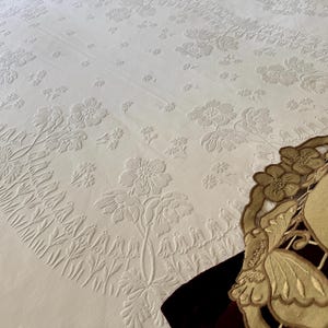 May include: A white bedspread featuring a raised, embossed floral pattern with intricate botanical motifs and a central oval design. In the lower right corner, a tan, circular decorative mat with cut-out flower and butterfly shapes rests.