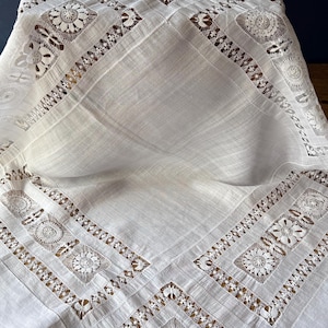 May include: White linen tablecloth with intricate lace and openwork detailing. The tablecloth features geometric patterns with floral and diamond motifs, creating an elegant and vintage aesthetic. The lace borders and center design add a touch of sophistication to the overall look.