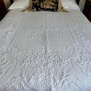 Op de afbeelding: Een witte sprei met een ingewikkeld verhoogd bloemen- en geometrisch patroon met een centraal stervormig motief. De stof heeft een getextureerd, reliëfachtig uiterlijk met symmetrische krulwerk- en botanische ontwerpen over het gehele oppervlak.