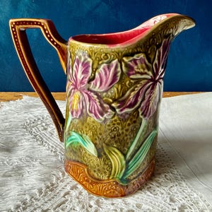 May include: A decorative ceramic jug with a handle. The jug features a floral design with purple irises on a green background. The handle is brown with gold accents, and the interior is red. The jug is approximately 23 cm tall.