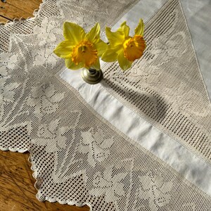 May include: A cream-coloured rectangular tablecloth featuring intricate lace borders with a floral pattern and a smooth central panel. Two bright yellow daffodils with orange centres are positioned in a small metal vase on the fabric surface.